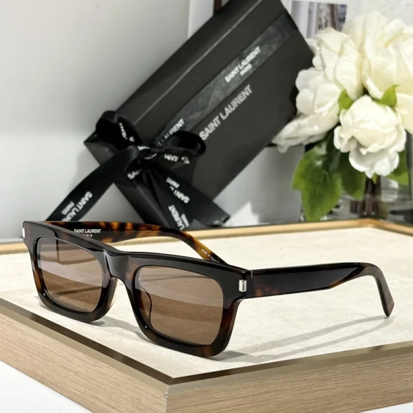 Front-facing Saint Laurent SL 461 Betty sunglasses placed on a white tabletop beside the branded case